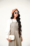 A woman wearing a beige pinafore dress with a white top, featuring a vintage-inspired neckline and wide straps.